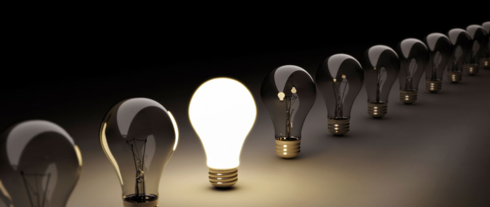 Fotografía de una fila de bombillas apagadas sobre un fondo negro, con una bombilla central brillando intensamente, simbolizando la innovación y el momento de encender la bombilla.